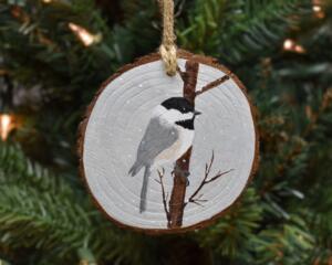 A handmade wood slice Christmas ornament with an acrylic painting of a chickadee in the snow at Cove Creek Photography.
